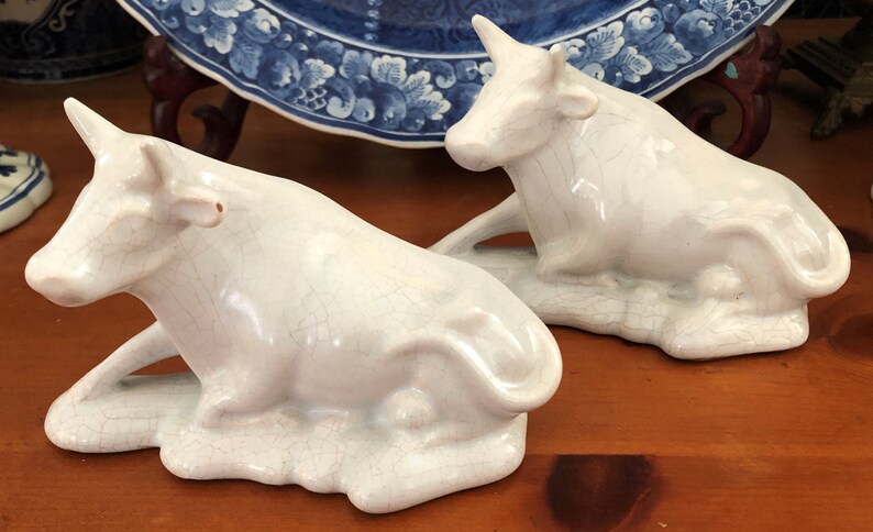 K&ouml;nnte beinhalten: Zwei wei&szlig;e Keramik-Kuhfiguren in entspannter Pose. Die Figuren haben eine Craquel&eacute;-Glasur und ruhen auf einem Sockel. Dekorationsartikel f&uuml;r ein Regal oder einen Tisch.