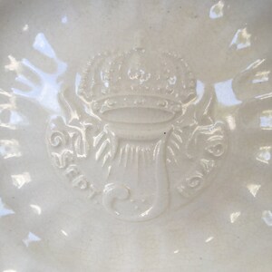 Puede incluir: Un plato de cer&aacute;mica blanca con un dise&ntilde;o en relieve de una corona y un motivo floral estilizado. El plato tiene un borde elevado y un acabado liso y brillante.