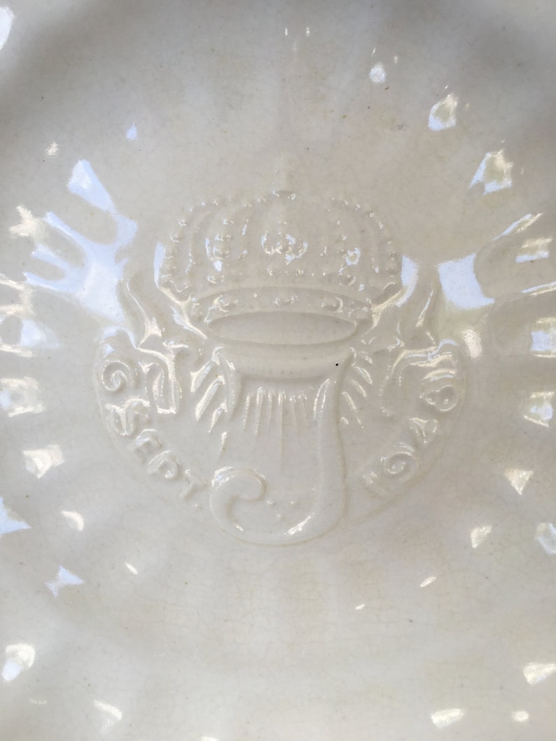 Puede incluir: Un plato de cer&aacute;mica blanca con un dise&ntilde;o en relieve de una corona y un motivo floral estilizado. El plato tiene un borde elevado y un acabado liso y brillante.