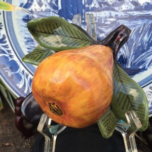 Puede incluir: Una escultura de cer&aacute;mica de pera con tonos naranja y amarillo, acentuada por hojas verdes y un tallo marr&oacute;n oscuro. La pera se presenta contra un plato con dise&ntilde;o azul y blanco, creando una naturaleza muerta decorativa.