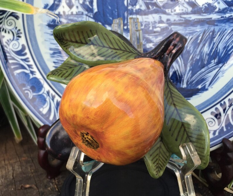 Puede incluir: Una escultura de cer&aacute;mica de pera con tonos naranja y amarillo, acentuada por hojas verdes y un tallo marr&oacute;n oscuro. La pera se presenta contra un plato con dise&ntilde;o azul y blanco, creando una naturaleza muerta decorativa.