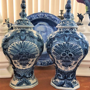 Może przedstawiać: Dwa pasujące niebiesko-białe ceramiczne wazony z pokrywkami, z misterne wzory kwiatowe i liściaste. Wazony mają pękaty kształt i są zdobione szczeg&oacute;łowym, ręcznie malowanym wzorem. Na tle widoczny jest pasujący talerz.
