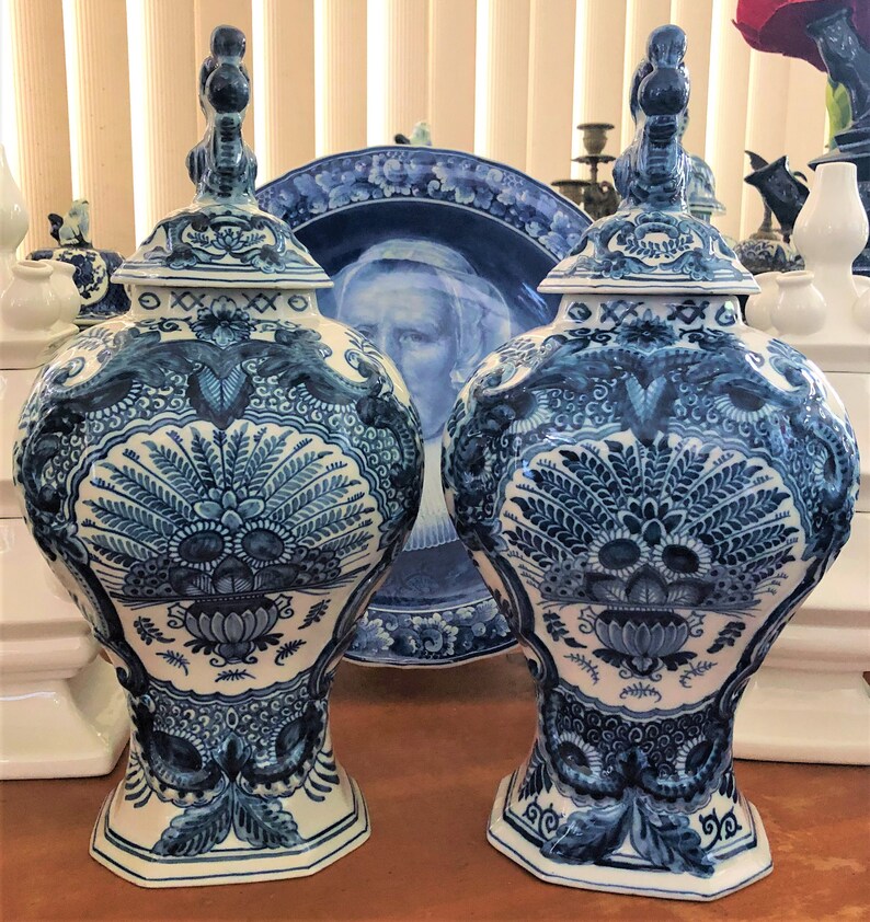 Może przedstawiać: Dwa pasujące niebiesko-białe ceramiczne wazony z pokrywkami, z misterne wzory kwiatowe i liściaste. Wazony mają pękaty kształt i są zdobione szczeg&oacute;łowym, ręcznie malowanym wzorem. Na tle widoczny jest pasujący talerz.