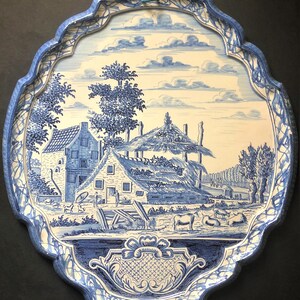 May include: Oval-shaped blue and white decorative tray with a rural landscape scene. The tray features a detailed illustration of a farmhouse, trees, and cows. The border has a scalloped edge with a blue and white pattern.