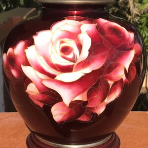 Puede incluir: Un jarr&oacute;n de cer&aacute;mica rojo con un dise&ntilde;o pintado a mano de una rosa blanca y rosa con detalles dorados. El jarr&oacute;n tiene un cuerpo redondo y una base de madera.