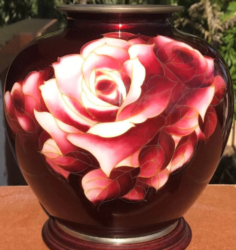 Puede incluir: Un jarr&oacute;n de cer&aacute;mica rojo con un dise&ntilde;o pintado a mano de una rosa blanca y rosa con detalles dorados. El jarr&oacute;n tiene un cuerpo redondo y una base de madera.