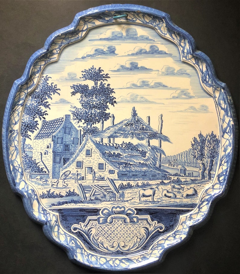 May include: Oval-shaped blue and white decorative tray with a rural landscape scene. The tray features a detailed illustration of a farmhouse, trees, and cows. The border has a scalloped edge with a blue and white pattern.