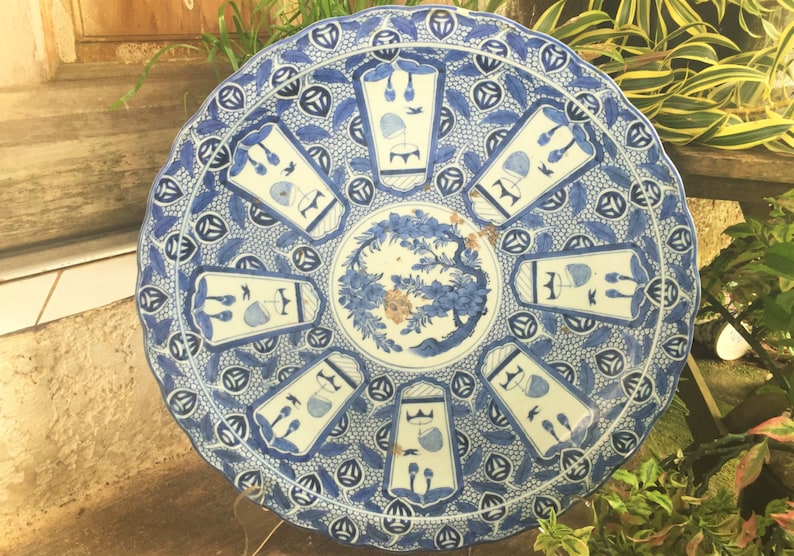 Op de afbeelding: Een grote, ronde, blauw-witte keramische plaat met een decoratief ontwerp. De plaat heeft een gegolfde rand en heeft een centraal bloemenmotief omringd door herhalende geometrische patronen.