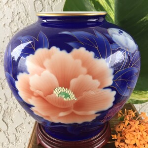 Puede incluir: Un jarr&oacute;n de cer&aacute;mica azul con un gran dise&ntilde;o de flor de peon&iacute;a color melocot&oacute;n. El jarr&oacute;n tiene un borde dorado y est&aacute; sobre un soporte de madera oscura. La flor tiene un centro verde y est&aacute; rodeada de hojas azules.