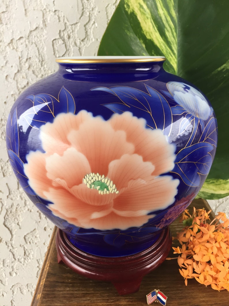 Puede incluir: Un jarr&oacute;n de cer&aacute;mica azul con un gran dise&ntilde;o de flor de peon&iacute;a color melocot&oacute;n. El jarr&oacute;n tiene un borde dorado y est&aacute; sobre un soporte de madera oscura. La flor tiene un centro verde y est&aacute; rodeada de hojas azules.