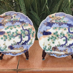 May include: Two decorative ceramic plates with a scalloped edge, each featuring a detailed landscape design. The plates have a white base with blue, gold, green, and peach accents. They are displayed on dark brown wooden stands.