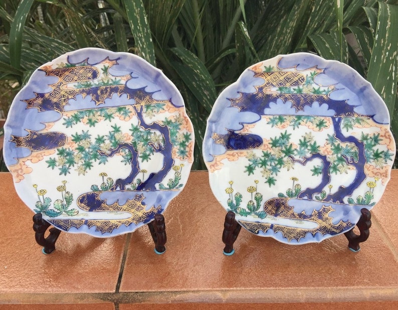 May include: Two decorative ceramic plates with a scalloped edge, each featuring a detailed landscape design. The plates have a white base with blue, gold, green, and peach accents. They are displayed on dark brown wooden stands.