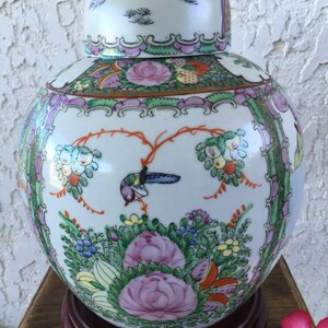 Op de afbeelding: Een witte porseleinen Chinese gemberpot met een deksel, versierd met bloemen- en vogelmotieven in tinten roze, groen en oranje. De pot staat op een houten voet.