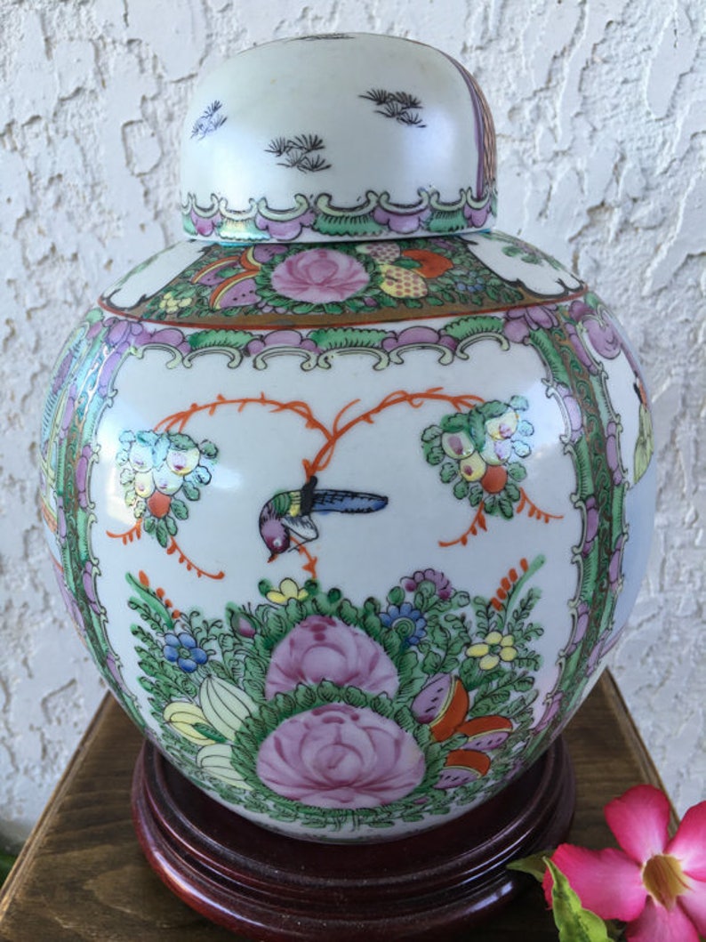 Op de afbeelding: Een witte porseleinen Chinese gemberpot met een deksel, versierd met bloemen- en vogelmotieven in tinten roze, groen en oranje. De pot staat op een houten voet.