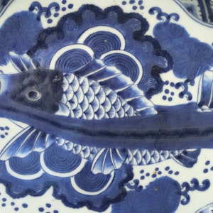 K&ouml;nnte beinhalten: Ein blau-wei&szlig;es Keramik-Tablett mit Fischmuster. Der Fisch ist in verschiedenen Blaut&ouml;nen mit detaillierten Schuppen und Flossen dargestellt. Das Tablett hat einen gewellten Rand und florale Muster, die einen klassischen, dekorativen Look erzeugen.