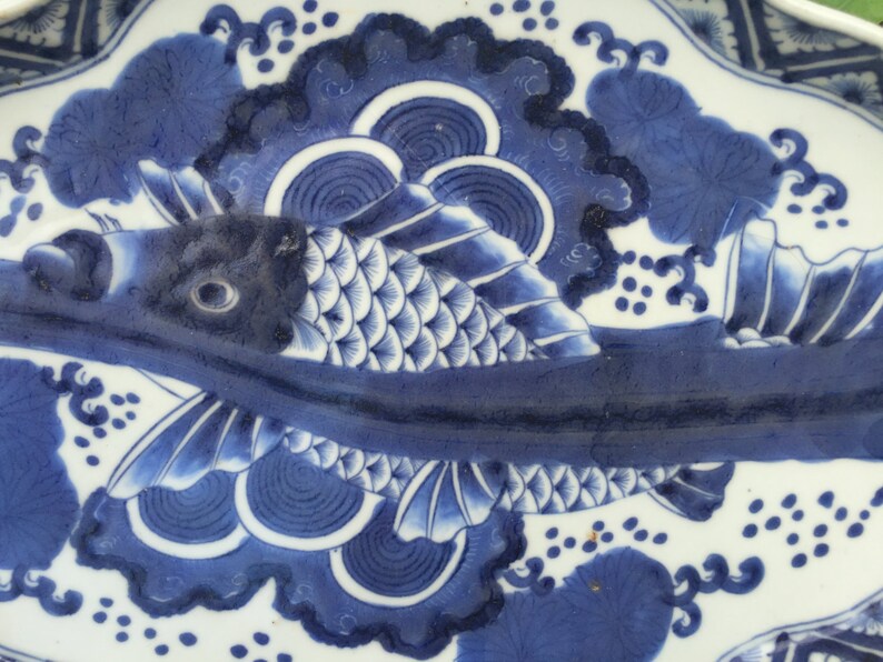 K&ouml;nnte beinhalten: Ein blau-wei&szlig;es Keramik-Tablett mit Fischmuster. Der Fisch ist in verschiedenen Blaut&ouml;nen mit detaillierten Schuppen und Flossen dargestellt. Das Tablett hat einen gewellten Rand und florale Muster, die einen klassischen, dekorativen Look erzeugen.