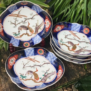 May include: A collection of decorative ceramic plates with a scalloped edge. The plates feature a blue and white patterned border, with a central design of birds and branches in shades of brown, orange, and green.