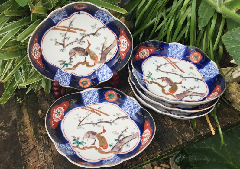 May include: A collection of decorative ceramic plates with a scalloped edge. The plates feature a blue and white patterned border, with a central design of birds and branches in shades of brown, orange, and green.