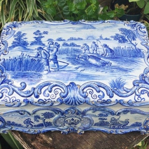 Puede incluir: Una caja de cer&aacute;mica azul y blanca con un dise&ntilde;o floral decorativo y una escena de personas trabajando en un campo. La caja tiene una tapa con bisagras y est&aacute; en buen estado.