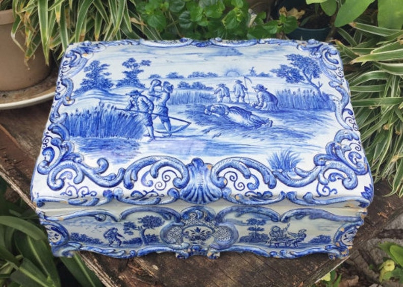 Puede incluir: Una caja de cer&aacute;mica azul y blanca con un dise&ntilde;o floral decorativo y una escena de personas trabajando en un campo. La caja tiene una tapa con bisagras y est&aacute; en buen estado.