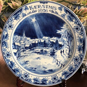 May include: A blue and white ceramic plate with a winter village scene. The plate features a border of holly and floral designs. The words "KERSTMIS 1956" are at the top. The plate is displayed on a wooden stand.