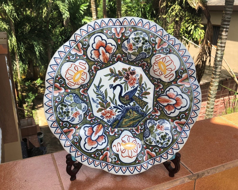 K&ouml;nnte beinhalten: Ein gro&szlig;er, runder, wei&szlig;er Teller aus Keramik mit einem gewellten Rand. Der Teller ist mit einem gr&uuml;nen, blauen und orangefarbenen Blumenmuster und einem Pfauenmotiv in der Mitte verziert.