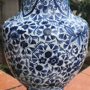 May include: A tall, ornate vase with a white and blue floral pattern. The ceramic vase features a wide body, a narrow neck, and a flared rim. The intricate design includes flowers, leaves, and swirling patterns. It sits on a wooden base.