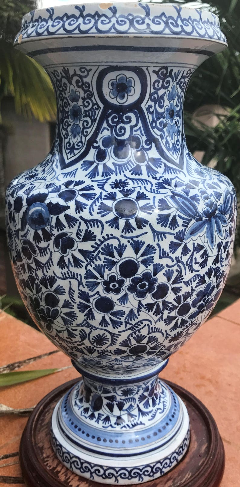 May include: A tall, ornate vase with a white and blue floral pattern. The ceramic vase features a wide body, a narrow neck, and a flared rim. The intricate design includes flowers, leaves, and swirling patterns. It sits on a wooden base.