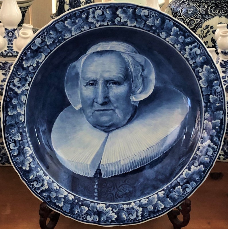Puede incluir: Un plato de Delftware azul y blanco con un retrato de una mujer que lleva un collar de encaje blanco y una gorra blanca. El plato est&aacute; decorado con un borde floral.