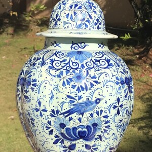 May include: A large, ornate ceramic vase with a lid, featuring a detailed blue and white floral and bird design. The vase is set on a dark wooden stand and is suitable for home decor.