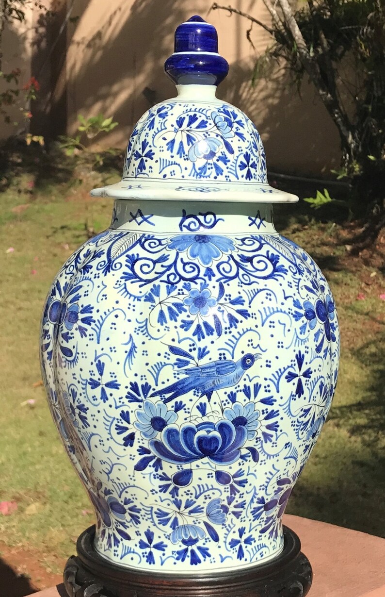 May include: A large, ornate ceramic vase with a lid, featuring a detailed blue and white floral and bird design. The vase is set on a dark wooden stand and is suitable for home decor.
