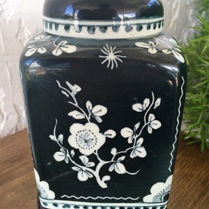 May include: A dark teal ceramic jar with a lid, featuring white floral and leaf designs. The jar has a square shape with rounded corners and sits on a black stand. The lid has a white and teal border.
