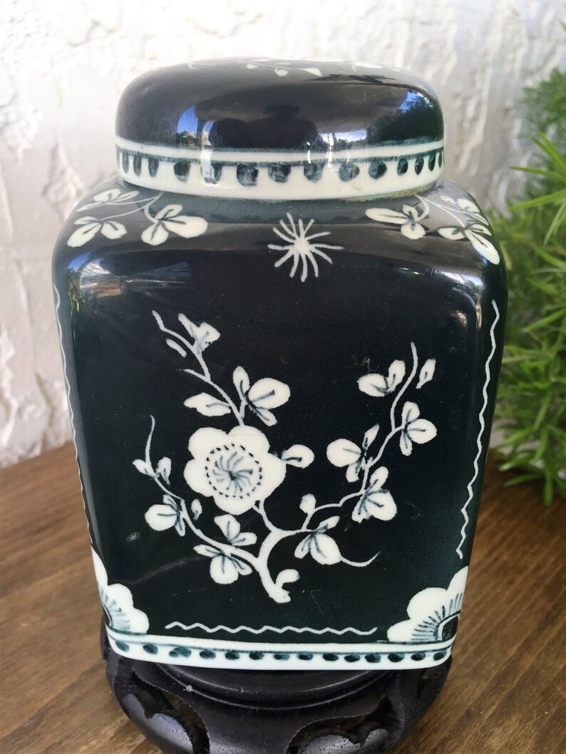 May include: A dark teal ceramic jar with a lid, featuring white floral and leaf designs. The jar has a square shape with rounded corners and sits on a black stand. The lid has a white and teal border.