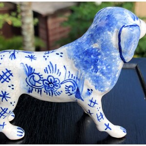 Puede incluir: Figura de perro de cer&aacute;mica en blanco y azul. El perro est&aacute; representado de pie, con orejas azules, espalda y marcas decorativas. Las marcas incluyen patrones florales y geom&eacute;tricos. Un objeto decorativo.
