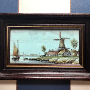 May include: A framed landscape painting depicting a Dutch windmill and a sailboat on a river. The artwork features a brown wooden frame with a gold inner edge. The scene includes a windmill, houses, trees, and a boat under a blue sky.