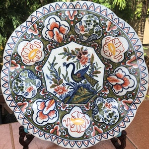 K&ouml;nnte beinhalten: Ein wei&szlig;er Teller aus Keramik mit gewelltem Rand zeigt ein farbenfrohes Blumen- und Pfauenmotiv. Der Teller ist mit blauer, gr&uuml;ner, roter und gelber Farbe verziert.