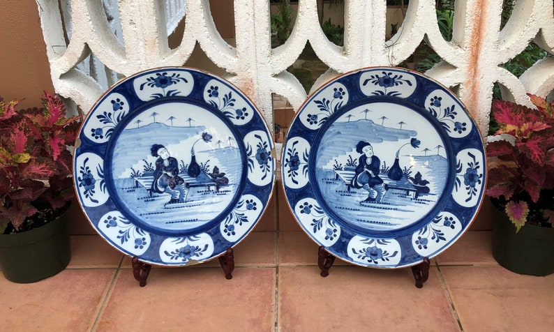 Puede incluir: Dos platos de cer&aacute;mica azul y blanco con un borde decorativo y una escena de una persona sentada en un banco en un paisaje. Los platos est&aacute;n sobre soportes de madera.