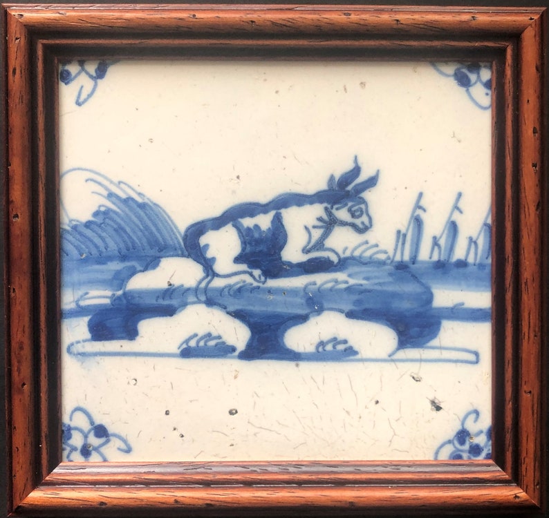 Puede incluir: Azulejo enmarcado con una ilustraci&oacute;n azul y blanca de un conejo o liebre sobre una roca. La obra de arte incluye elementos estilizados de agua y plantas. El azulejo est&aacute; enmarcado en un marco de madera marr&oacute;n oscuro.