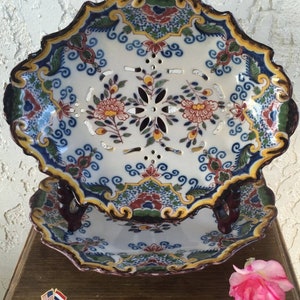 Puede incluir: Dos platos vintage de Delftware holandeses con motivos florales azules, blancos, verdes, amarillos y rojos. El plato superior tiene un dise&ntilde;o calado decorativo y est&aacute; apilado sobre un plato s&oacute;lido. Ambos platos tienen bordes festoneados y est&aacute;n decorados con un borde marr&oacute;n.