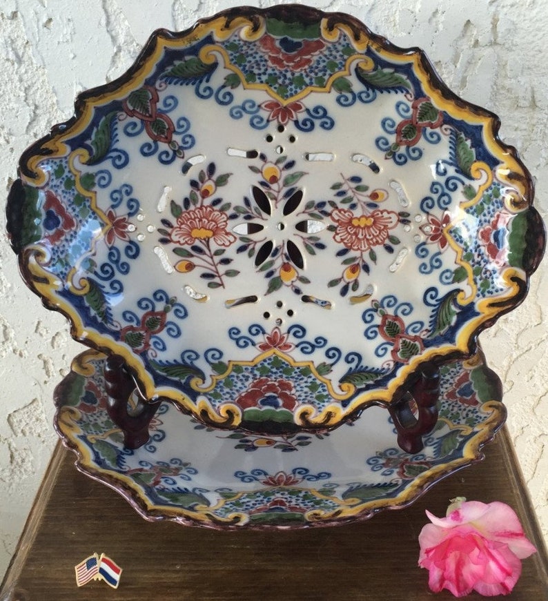 Puede incluir: Dos platos vintage de Delftware holandeses con motivos florales azules, blancos, verdes, amarillos y rojos. El plato superior tiene un dise&ntilde;o calado decorativo y est&aacute; apilado sobre un plato s&oacute;lido. Ambos platos tienen bordes festoneados y est&aacute;n decorados con un borde marr&oacute;n.