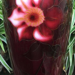 Puede incluir: Un jarr&oacute;n rojo oscuro con un dise&ntilde;o de flores blancas y doradas. El jarr&oacute;n tiene un borde plateado y se asienta sobre una base de madera negra.