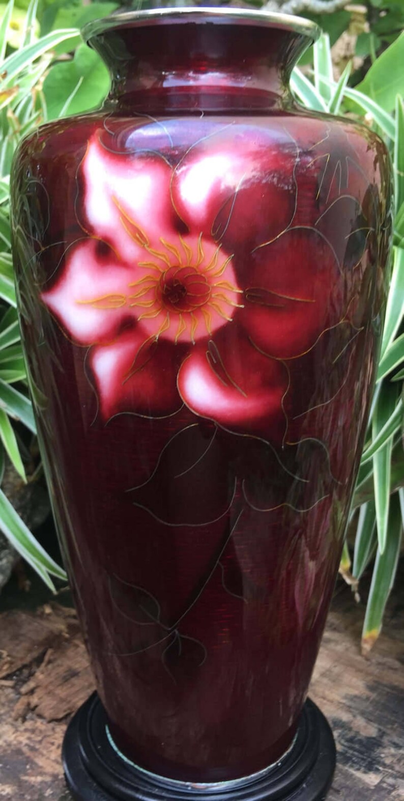 Puede incluir: Un jarr&oacute;n rojo oscuro con un dise&ntilde;o de flores blancas y doradas. El jarr&oacute;n tiene un borde plateado y se asienta sobre una base de madera negra.