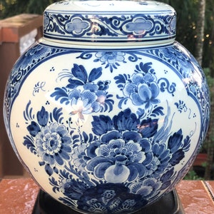 May include: A white and blue ceramic jar with a lid. The jar features a hand-painted floral design in shades of blue. The lid and body are adorned with intricate patterns. The jar is displayed on a black stand.