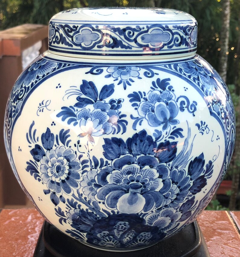 May include: A white and blue ceramic jar with a lid. The jar features a hand-painted floral design in shades of blue. The lid and body are adorned with intricate patterns. The jar is displayed on a black stand.