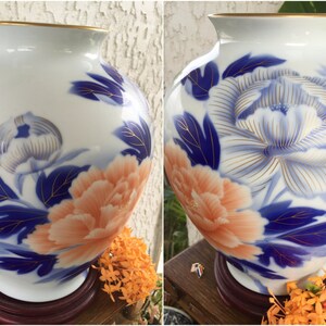 Puede incluir: Jarr&oacute;n de cer&aacute;mica blanca con borde dorado, con un dise&ntilde;o floral en melocot&oacute;n, azul y dorado. El jarr&oacute;n tiene forma redonda y se apoya sobre una base de madera. El dise&ntilde;o floral incluye una peon&iacute;a.