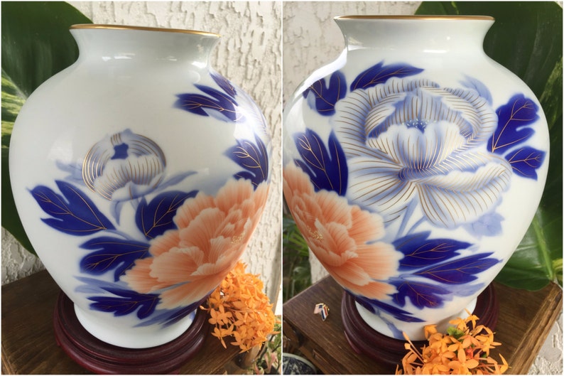 Puede incluir: Jarr&oacute;n de cer&aacute;mica blanca con borde dorado, con un dise&ntilde;o floral en melocot&oacute;n, azul y dorado. El jarr&oacute;n tiene forma redonda y se apoya sobre una base de madera. El dise&ntilde;o floral incluye una peon&iacute;a.