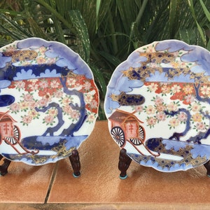 May include: Two decorative ceramic plates with scalloped edges, featuring a Japanese-style design. The plates showcase a scene with a tree, flowers, and a carriage, with blue, red, and gold accents. Each plate is displayed on a wooden stand.