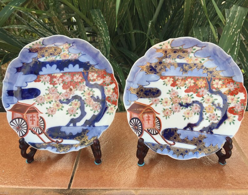 May include: Two decorative ceramic plates with scalloped edges, featuring a Japanese-style design. The plates showcase a scene with a tree, flowers, and a carriage, with blue, red, and gold accents. Each plate is displayed on a wooden stand.