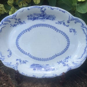 May include: An oval white ceramic platter featuring a blue transferware pattern. The design includes a central decorative border, intricate scrollwork along the scalloped edges, and detailed landscape scenes depicting a windmill in blue ink.