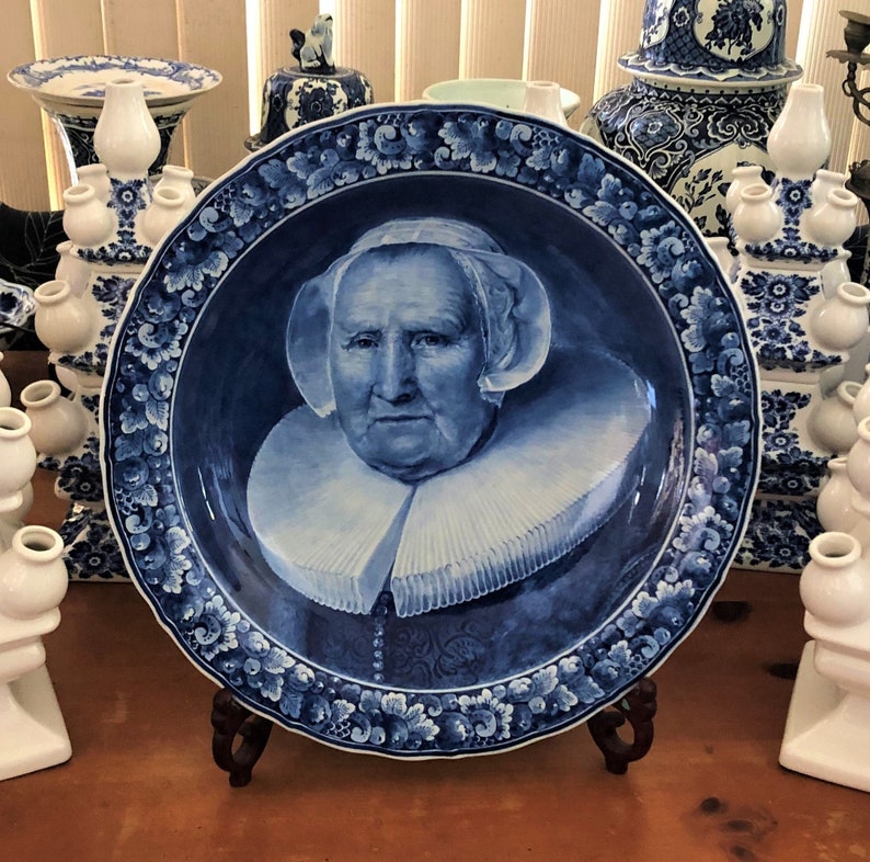 May include: A blue and white Delftware plate featuring a portrait of a woman wearing a white lace collar and a white cap. The plate has a decorative border of blue flowers and leaves.